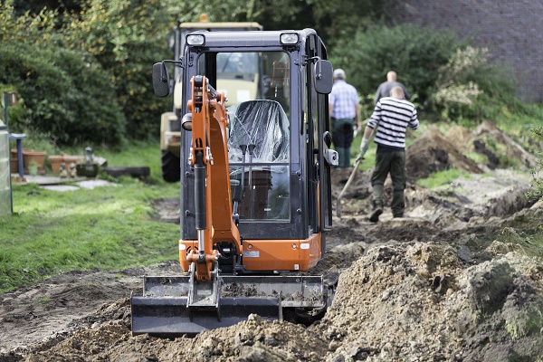 A digger vehicle excavates, with workmen in the background